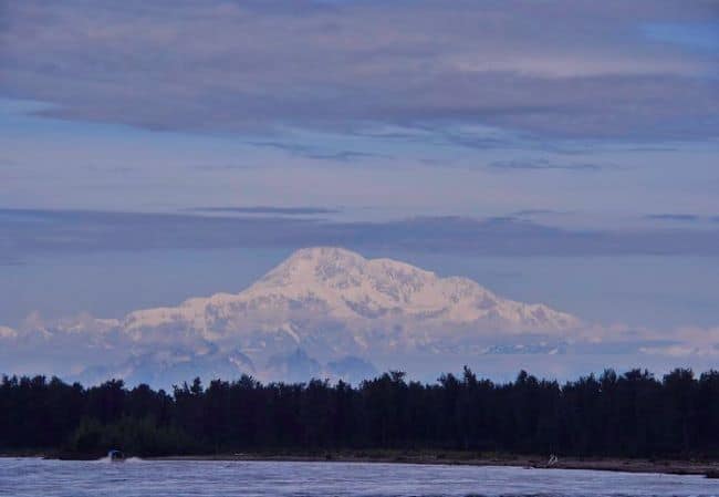 Snow-capped  Denali mountain with trees and a river in the foreground under a cloudy sky.