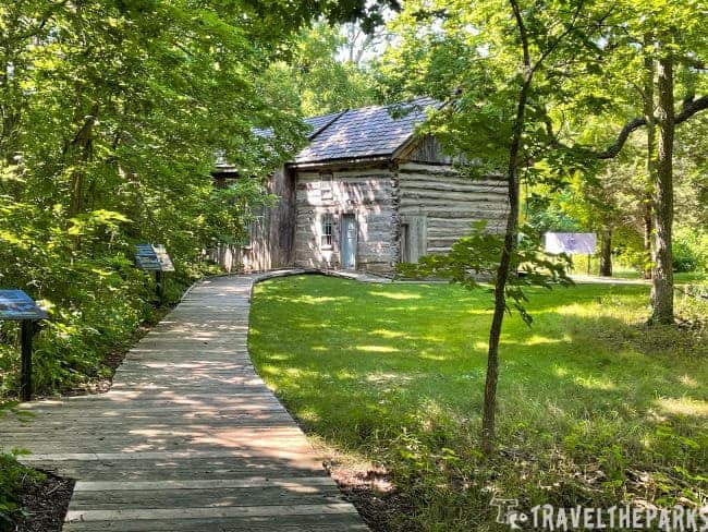A wooden boardwalk leads through a green forest to a log cabin.