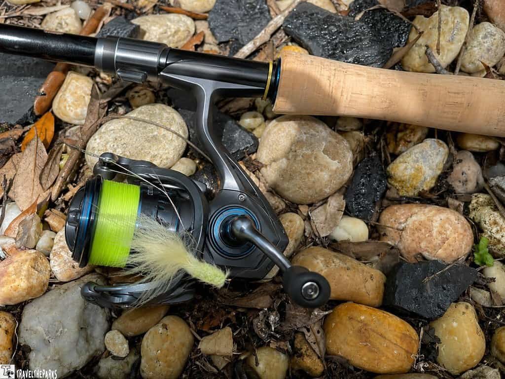 Spinning fishing rod and reel with neon-green line and a pale artificial lure lying on rocks and leaves.