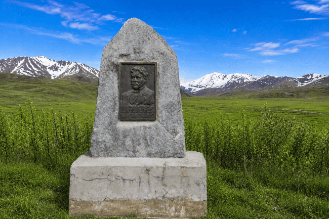 Gray stone monument with a bronze plaque of a man's bust in a green landscape with snow-capped mountains and a blue sky.