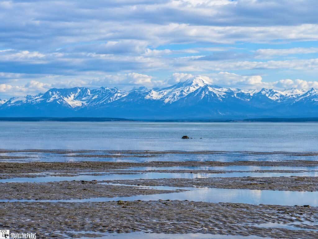 Snow-capped mountains rise beyond a calm bay and muddy tidal flats beneath a cloudy blue sky near Kenai.