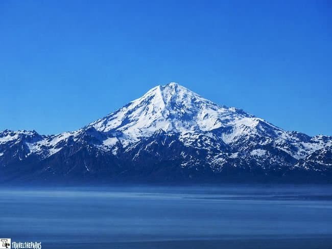 Snow-covered mountain range reflected across calm blue water beneath a clear sky.