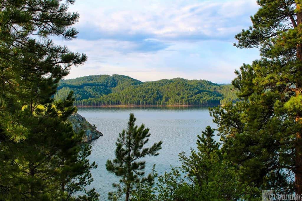 A calm lake surrounded by forested hills under a partly cloudy sky, framed by evergreen trees.