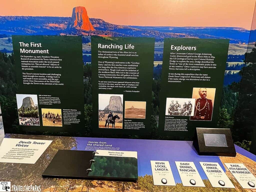 a museum exhibit with panels in front of a photo backdrop featuring Devils Tower, an iconic geological formation against an orange-hued sky at sunset. The exhibit consists of three main informational panels: "The First Monument," "Ranching Life," and "Explorers." Each panel contains text and images related to its topic. The panels are visually similar, with a dark green background and white text. Below these, a flat display surface has materials titled "Devils Tower Voices," which include tactile and printed booklets and tags with names and Braille. There is also a small digital display off to the right.