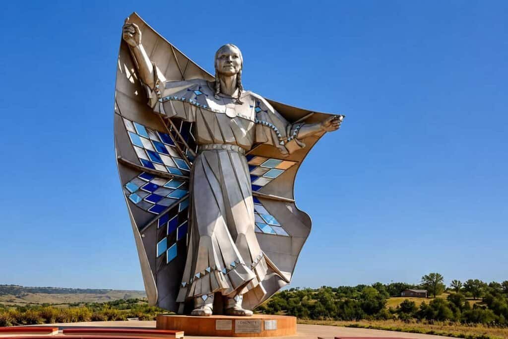 Dignity Statue: A metallic statue of a Native American woman stands with arms raised, wearing traditional clothing adorned with blue and silver patterns.

