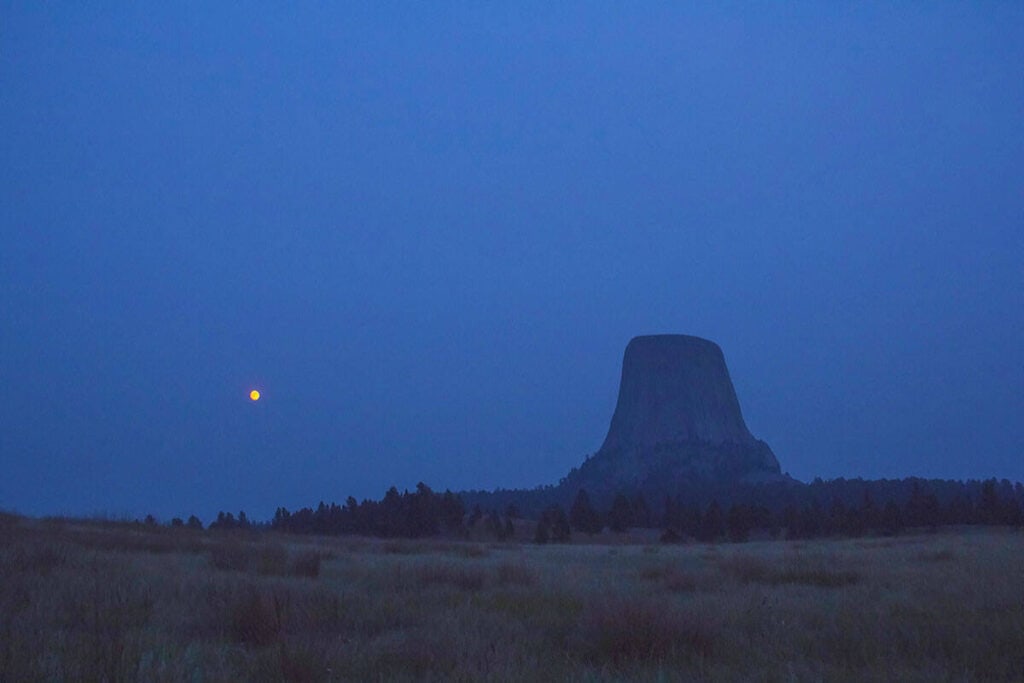 View of Devils Tower at dusk with a glowing orange moon in a deep blue sky.