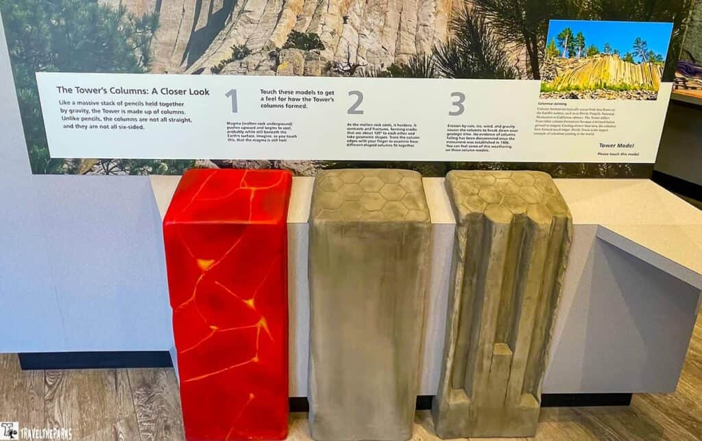 a display panel at an informational exhibit about the formation of geological columns. The top section of the image contains a descriptive panel with text and images. Below it, three models are presented to illustrate different stages of column formation. The left model is vibrant red with glowing orange vein-like patterns, resembling molten rock. The middle model is a grayish color, portraying a hexagonal column top texture. The right model is also gray, showcasing elongated and slightly eroded columns, simulating natural weathering. All models sit on a light-colored platform.