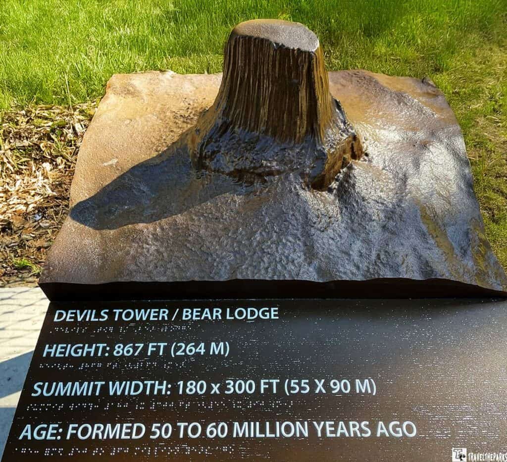 a detailed model of Devils Tower, a notable geographical formation. The model is a three-dimensional representation, showing the tower's vertical columns rising from its base, mimicking its natural form. It is mounted on a rectangular slab, placed on a grassy area, with sunlight casting shadows. The surface of the model is textured, reflecting the rock's uneven and striated appearance.
In front of the model is a black informational plaque with white text and braille dots, providing geological and dimensional details about Devils Tower, like its height, width, and age.