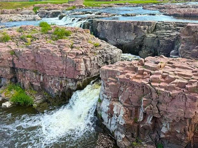 View of Sioux Falls with cascading water over reddish-brown rocks and surrounding greenery.