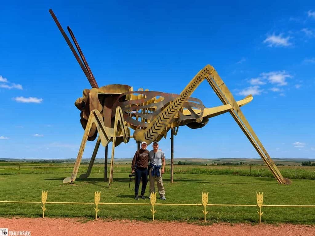 Large metal sculpture of a grasshopper with two people posing in front, set against a blue sky and green grass. A decorative yellow fence runs along the foreground.

