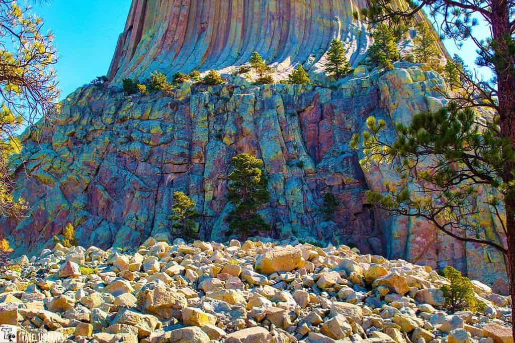 Base of a large multicolored rock formation with trees and boulders in the foreground.