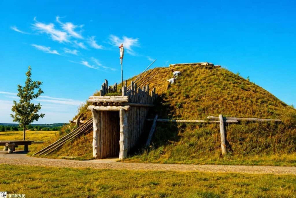 A traditional Hidatsa Earth lodge at Knife Village, featuring a grassy hill roof, log entrance, and a flagpole, set against a bright blue sky.

