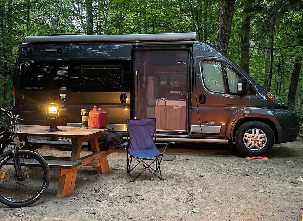 RV parked at a wooded campsite with a picnic table, lantern, and folding chair.