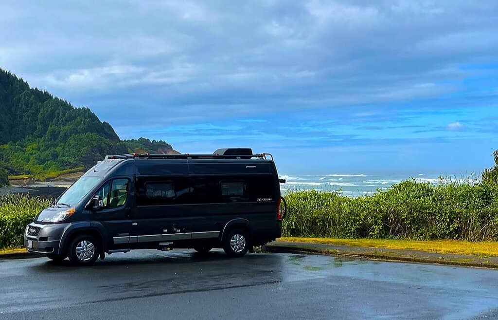 Black RV parked in front of coastal landscape with green hills and ocean in the background.