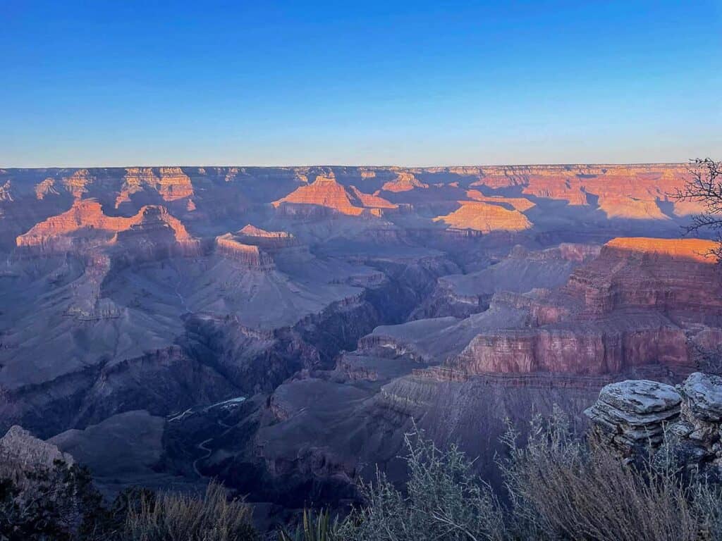 Sunset view of the Grand Canyon with orange and red hues on rock formations, under a clear blue sky.