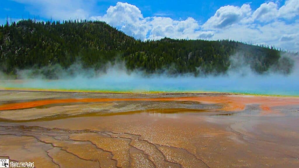 Colorful geothermal hot spring with steam, surrounded by a forested hill and cloudy sky.