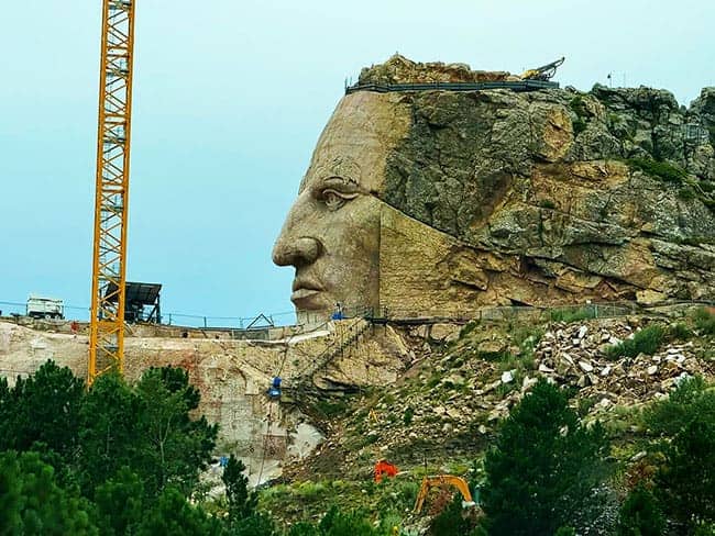 Profile of a Native American face carved into a rocky mountain, with construction equipment nearby.