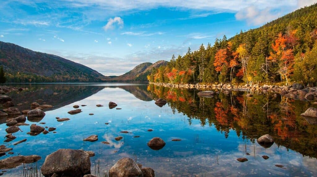 A tranquil lake in Acadia National Park reflecting autumn-colored trees and hills under a blue sky.