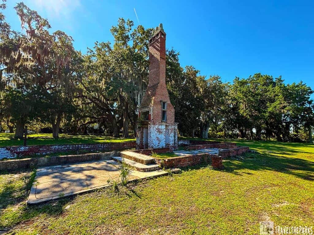 Old brick chimney remains with trees and hanging moss under a clear blue sky.

