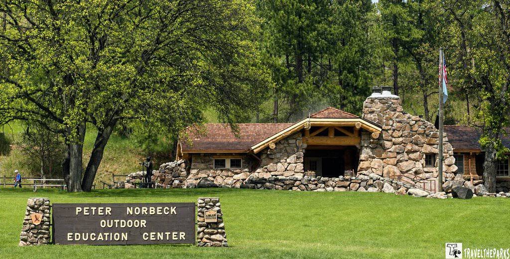 Outdoor scene with a stone building, grassy lawn, trees, and a sign reading "Peter Norbeck Outdoor Education Center."