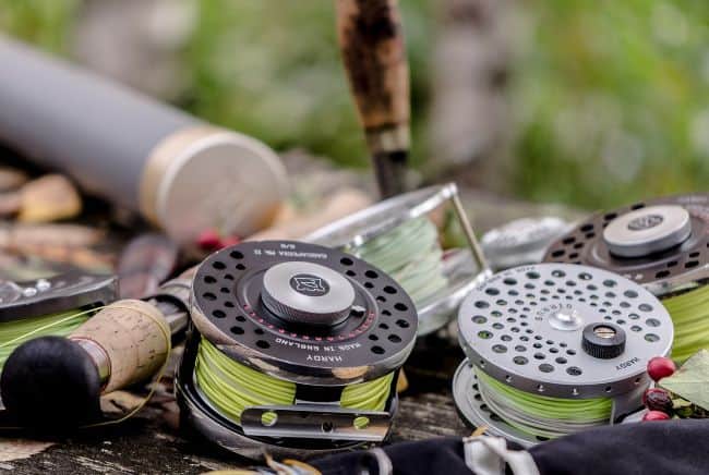 A close-up of fly fishing reels and a fishing rod on a wooden surface, showcasing bright green fishing line and scattered leaves and berries.