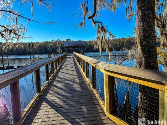 Wooden boardwalk leading to a pavilion over calm water, surrounded by trees with hanging Spanish moss.