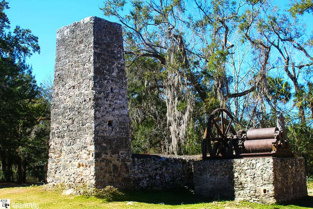 ruins of the Yulee Sugar Mill set in a lush, green landscape. Dominating the foreground is a rectangular stone structure, visibly aged with weathered grey and white stones. Atop this structure, a large, rusty iron wheel and gears are partially visible. To the right, there is a tall, rectangular stone chimney of similar construction. Surrounding the ruins are grassy areas with scattered small stones and patches of fallen leaves. Tall trees with sprawling, curved branches and draped with Spanish moss frame the scene, casting dappled shadows on the ground. The sky above is bright blue with a few white clouds, adding a vivid contrast to the earthy tones of the mill and the green foliage.