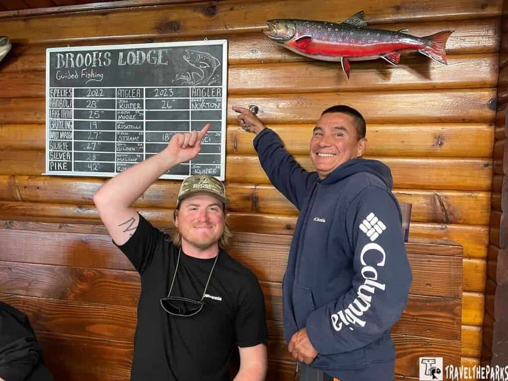 The Brooks Lodge Fishing Achievement Board: Two people in front of a wooden wall, with a chalkboard and a mounted fish above them.