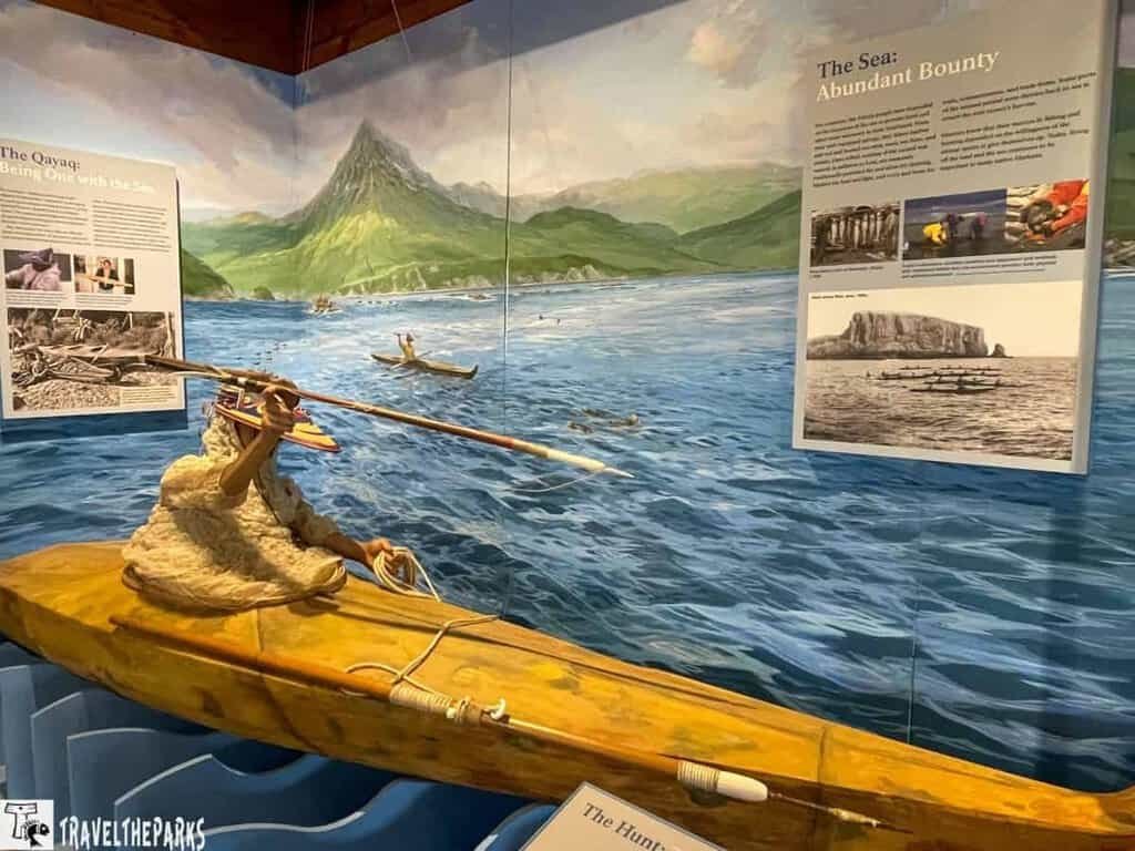 A lifelike figure of a paddler in a kayak, dressed in a fur-like garment, is shown against a colorful ocean backdrop with mountains. Informational panels about kayaks and marine life are nearby.

