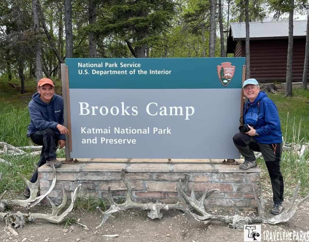 People stading in front of the Brooks Camp Sign. Antlers are scattered in front of the sign with trees int he background.