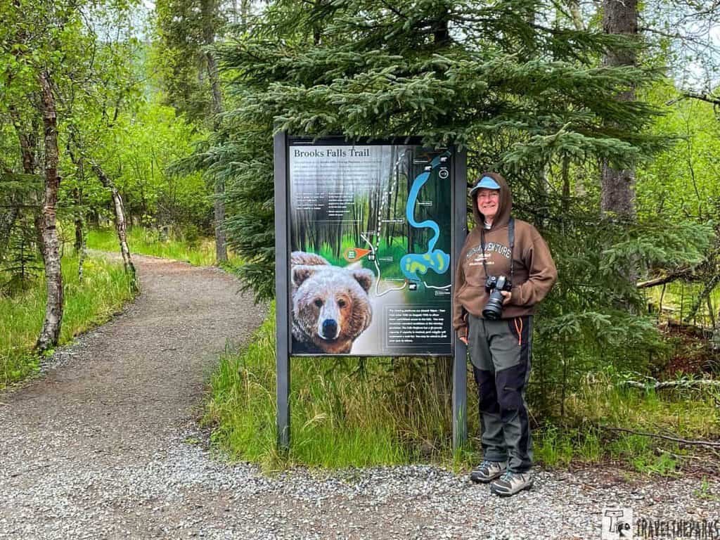 Brooks Trail Sign with women in front: Person standing next to a Brooks Falls Trail sign with a bear image in a forested area.