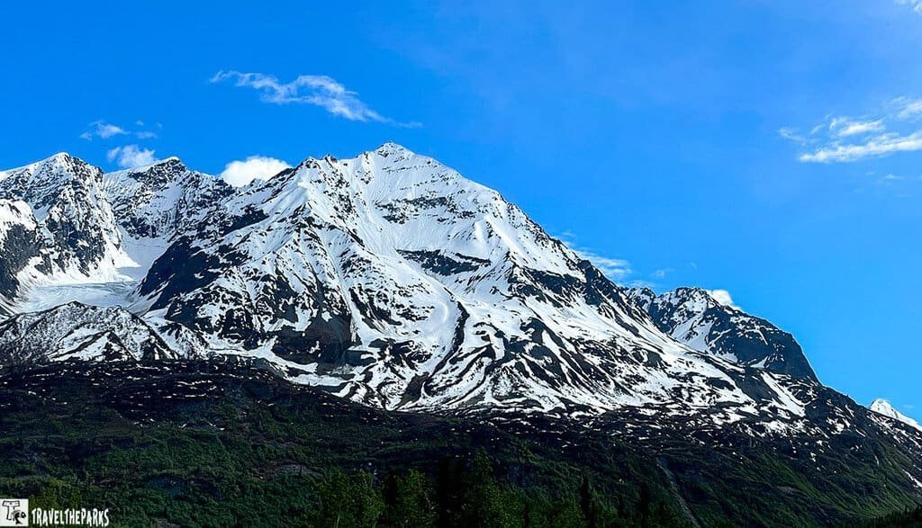 A scenic view of the Chugach Mountains with snow-capped peaks against a blue sky, featuring rocky slopes and green vegetation in the foreground.