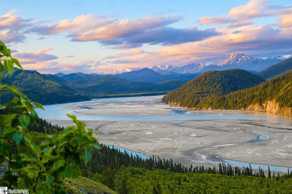 A scenic view of the Copper River along McCarthy Road, featuring flowing water, sandy riverbanks, green mountains, and a pastel-colored sky during sunrise or sunset.