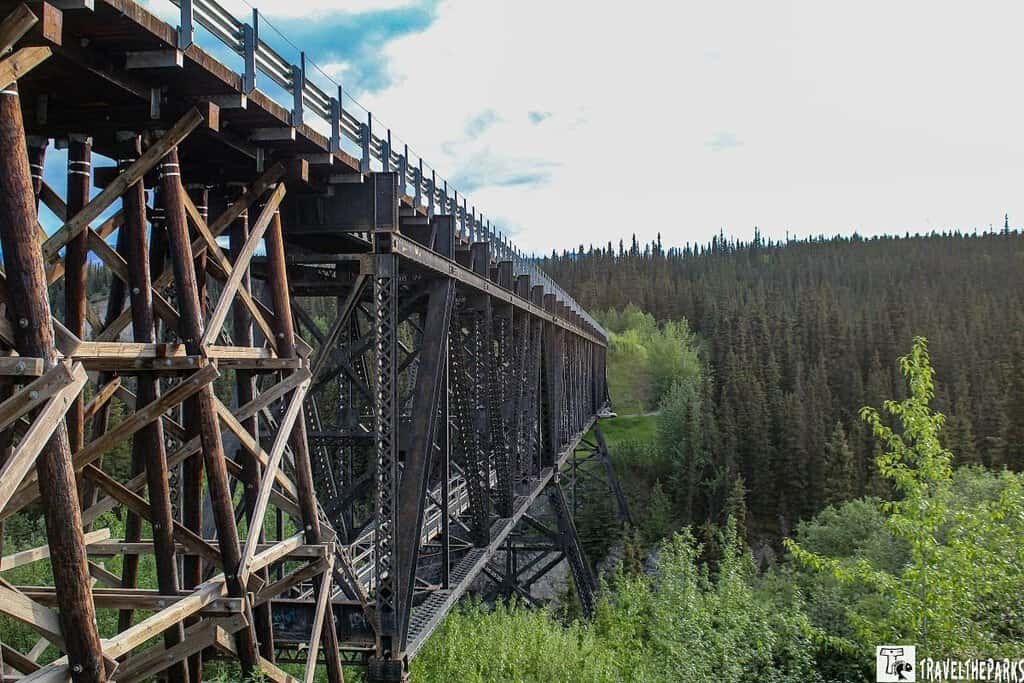 Kuskulana River Bridge: Wooden trestle bridge over a forested landscape under a cloudy sky in Wrangell St Elias NP.