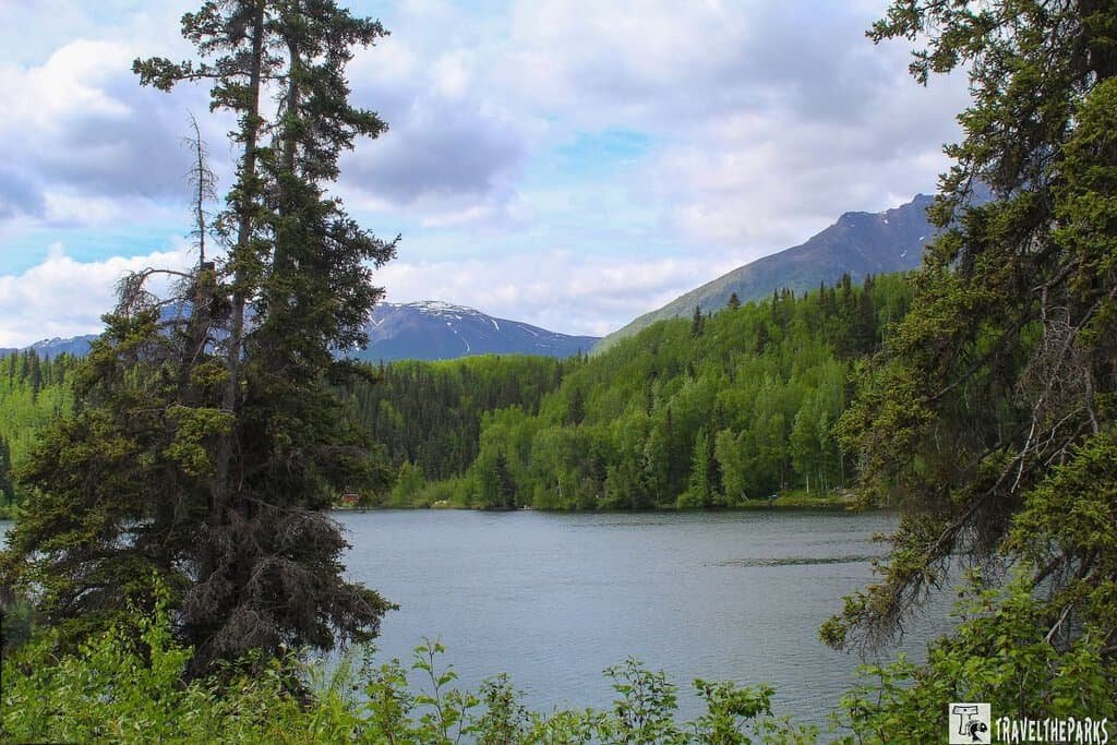 Scenic view of a calm Long Lake surrounded by lush green mountains and evergreen trees under a cloudy sky.