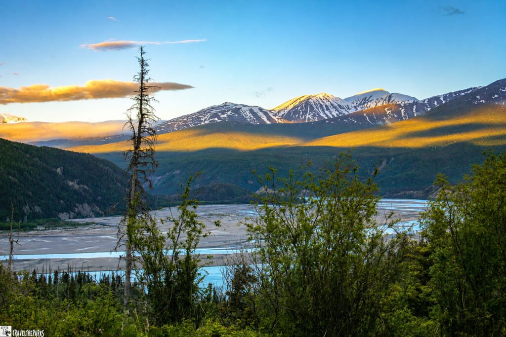 A scenic view of the Copper River with rugged mountains in the background, green bushes in the foreground, a winding river, and a sunset casting golden light on the landscape.