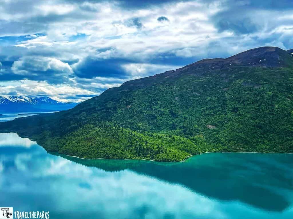 Flying over Naknek Lake to brooks Camp: A serene landscape with a reflective body of water, a lush green mountain, and distant snow-capped peaks under a cloudy sky.