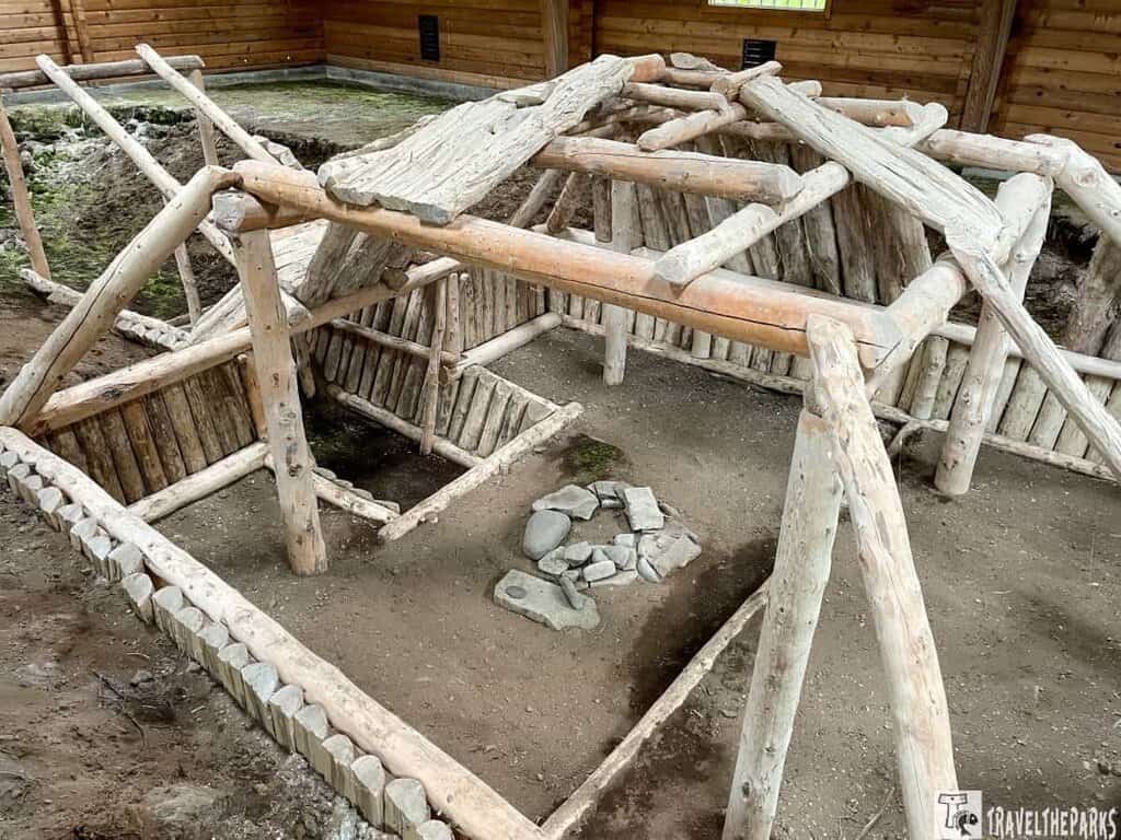 Reconstructed ancient wooden structure inside a sheltered area, featuring log beams and a central stone hearth.