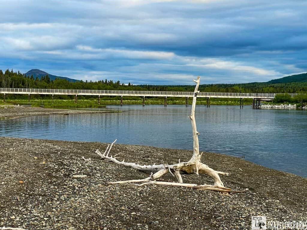 Lower River elevate boardwalk at Brooks Camp: A river with a gravel shoreline, driftwood, and a wooden pedestrian bridge under a cloudy sky.