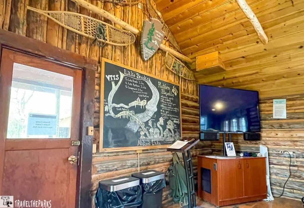 Bear school orientation in the Brooks Camp Visitor Center: Rustic cabin interior with a wooden door, chalkboard with a landscape drawing, snowshoe, mounted television, and wooden cabinetry.