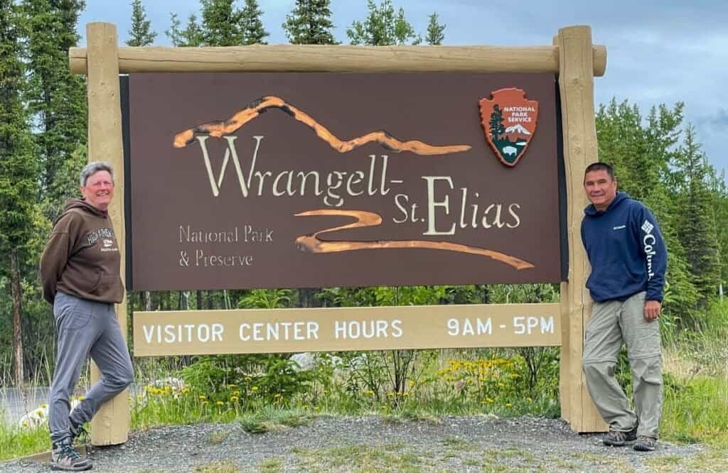 Two people standing by the Wrangell-St. Elias National Park sign with pine trees in the background.