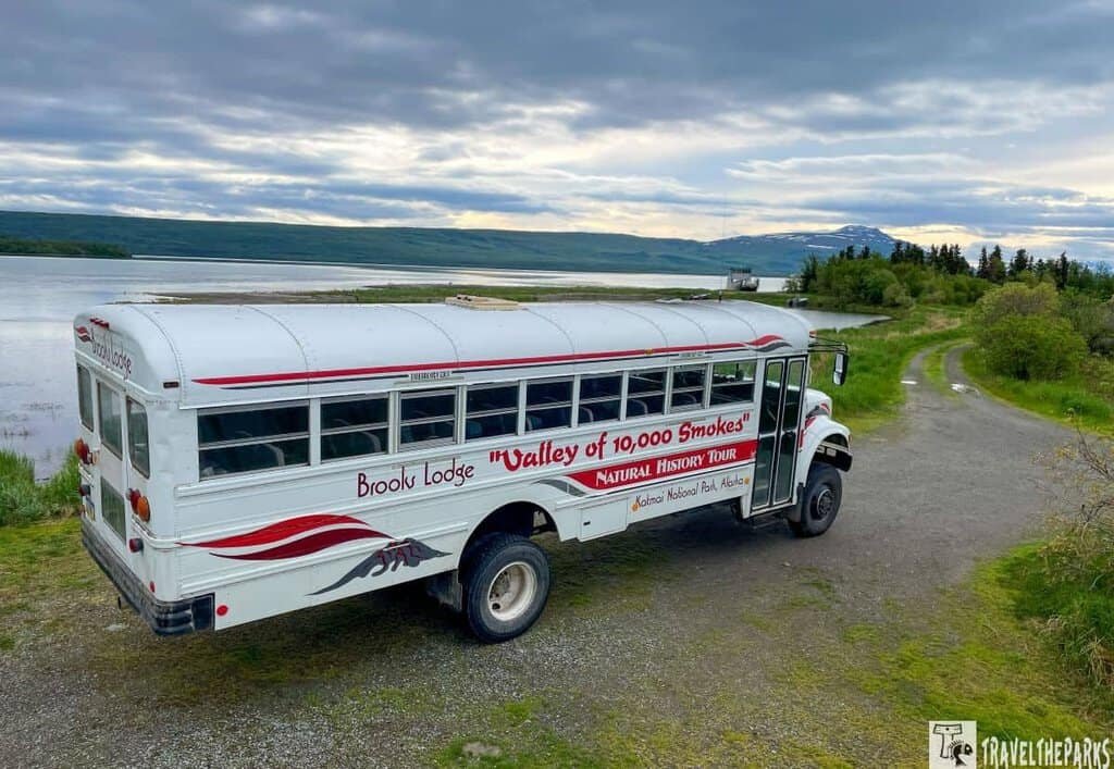 Valley of Ten Thousand Smokes Tour Bus-Katmai. White bus on a gravel road near a large body of water with hills and cloudy sky.
