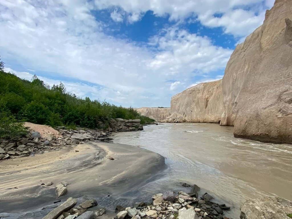 A winding brown river flows through the Valley of 10,000 Smokes, flanked by steep light-colored cliffs and green vegetation along the banks.