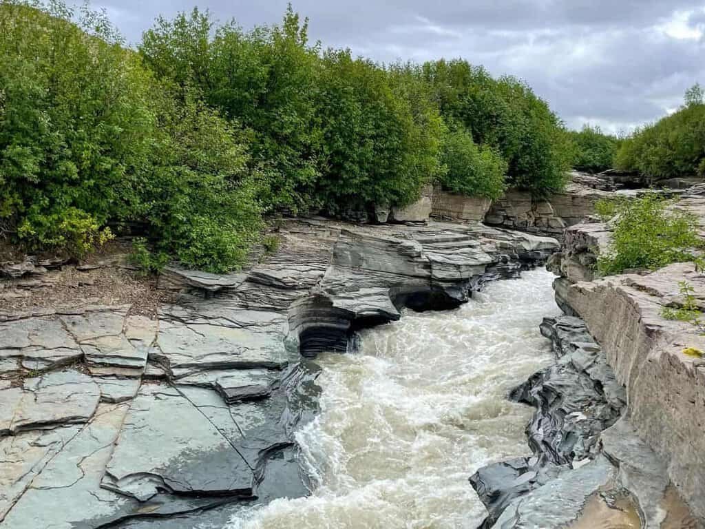 Rugged riverbanks with smooth, layered rocks surround a rushing river, framed by dense greenery on a cloudy day.

