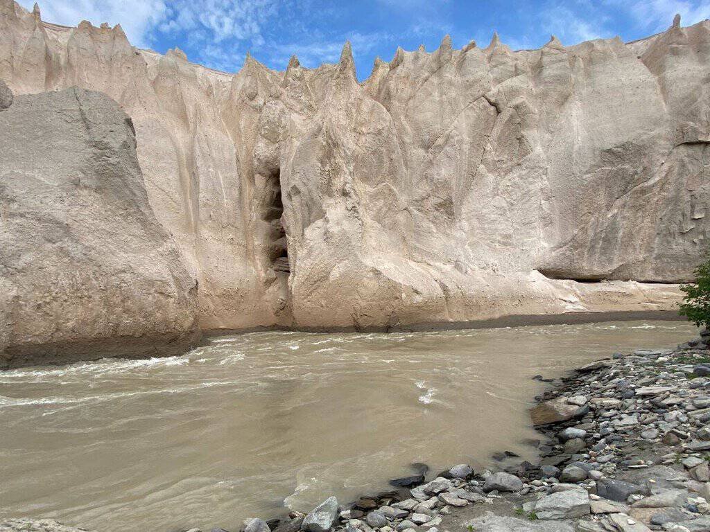 A landscape image of the Valley of Ten Thousand Smokes showing beige cliffs with sharp peaks beside a muddy river and rocky banks.

