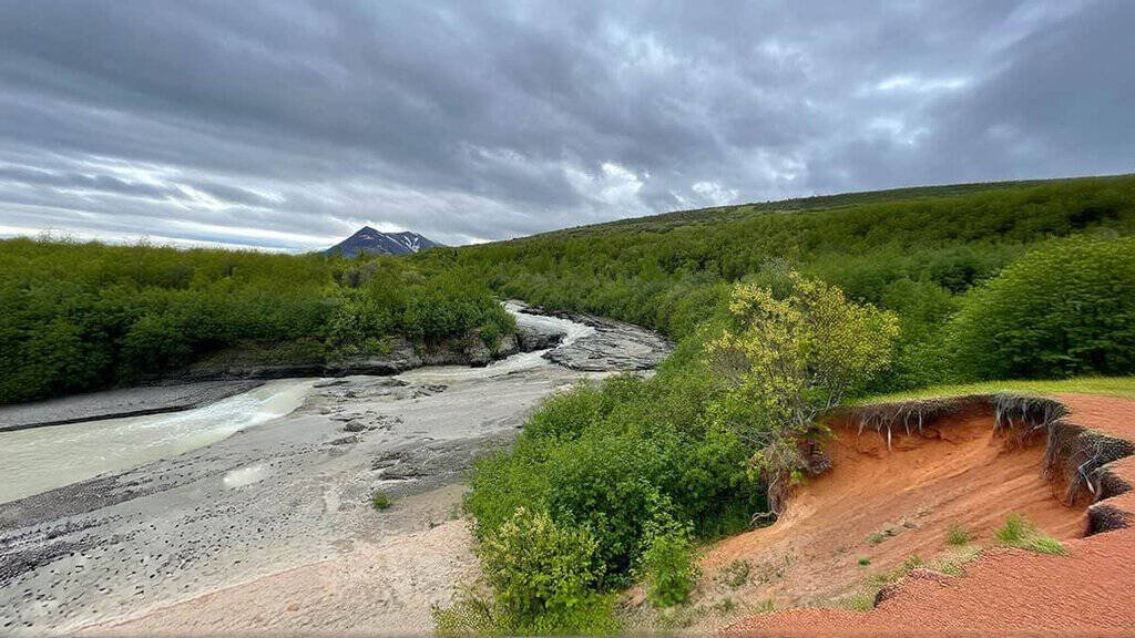 Knife River Valley of 10,000 Smokes: A river winding through a green, forested valley with a mountain and cloudy skies in the background.