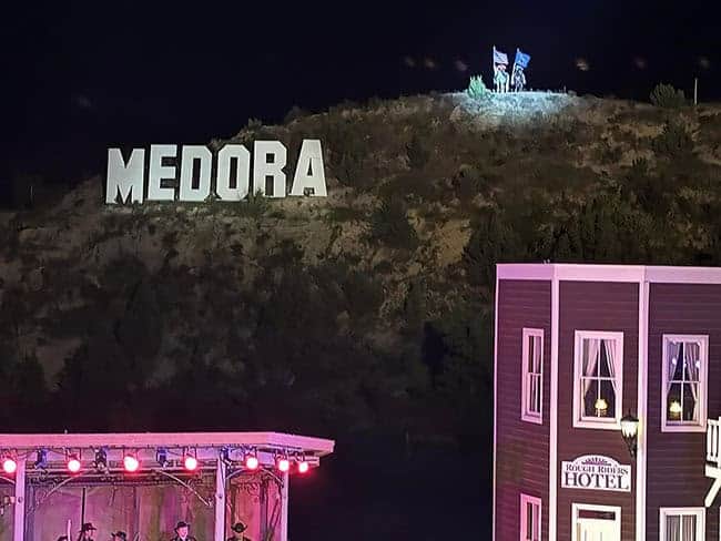 Nighttime scene featuring a "MEDORA" sign on a hillside, with two individuals holding flags above it. In the foreground, the "ROUGH RIDERS HOTEL" is visible, lit with pink and red lights.