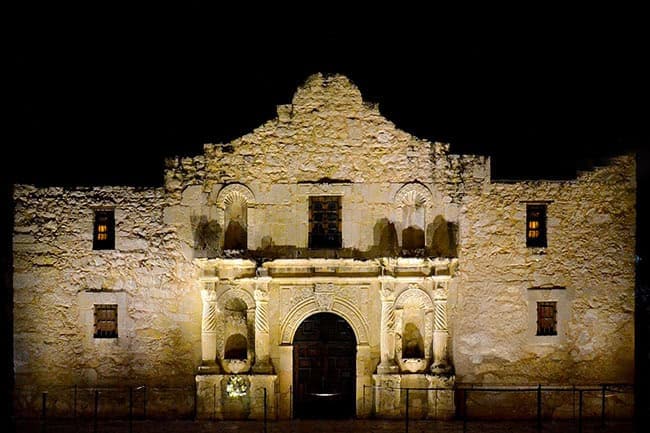 the Alamo's front facade at night. The building is constructed from light-colored stone, illuminated by spotlights that highlight its architectural details against the dark sky. The facade is symmetrical, featuring a prominent arched doorway in the center, flanked by two columns and intricate carvings. Above the doorway is a small rectangular window. The upper section of the facade has a scalloped top edge. Two small windows with visible interiors are positioned symmetrically on either side of the facade. The building's texture is rough, adding a historical, weathered appearance.