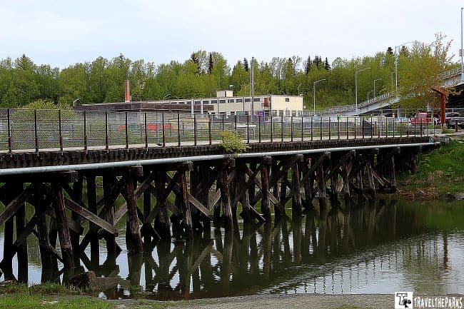 Wooden bridge with crisscross pylons over water, surrounded by green trees and a building in the background. Ship Creek in Anchorage Alaska