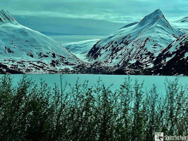 Snowy mountains near Portage Lake with turquoise water and green shrubs in the foreground.